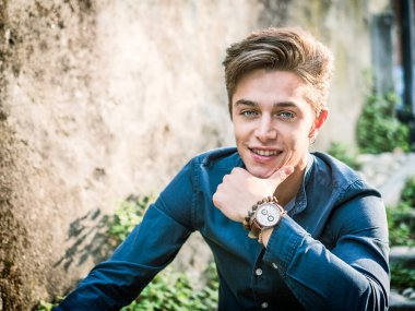 Avrupa 'nın kentsel ortamında, güneşli bir günde oturmuş mutlu bir şekilde gülümseyen kameraya bakan yakışıklı, zarif bir genç.