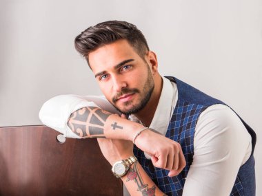 Stüdyoda şık bir yelek ve beyaz gömlek giyen genç adam kameraya bakıyor.