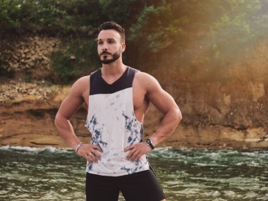 Nehir kenarındaki yakışıklı atletik genç adam, elleri kalçasında, başka tarafa bakıyor.