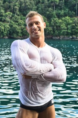 Attractive young bodybuilder by the sea with wet shirt on, smiling expression, looking at camera with arms crossed on chest