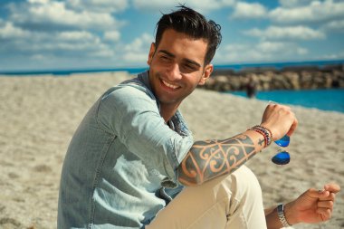 Kumsaldaki yakışıklı genç adam, gülümsüyor, kumda oturuyor, kot gömlek ve beyaz pantolon giyiyor.