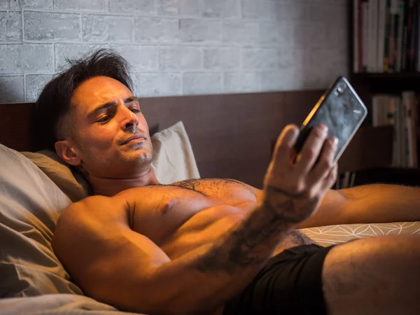Attractive athletic young man using cell phone to take selfie photo or watching something on mobile, while laying shirtless on bed, shirtless