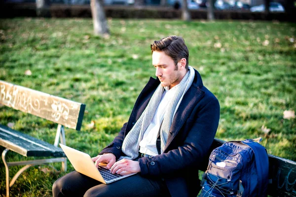 真剣な表情で彼のラップトップ コンピューターに情報を入力する都市公園における屋外での作業 木製のベンチの上に座ってハンサムなエレガントなビジネスマン — ストック写真