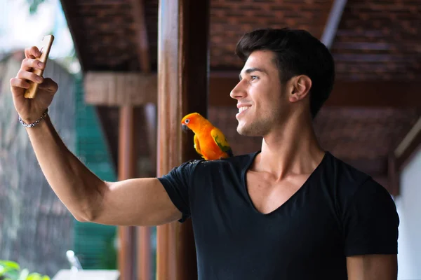 Uśmiechnięty Przystojniak Robiący Zdjęcie Papugi Siedzącej Ramieniu Strzał Poziomie Wewnątrz — Zdjęcie stockowe