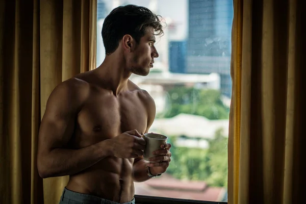 Sexy handsome young man standing shirtless in his bedroom drinking a cup of coffee or tea next to window curtains