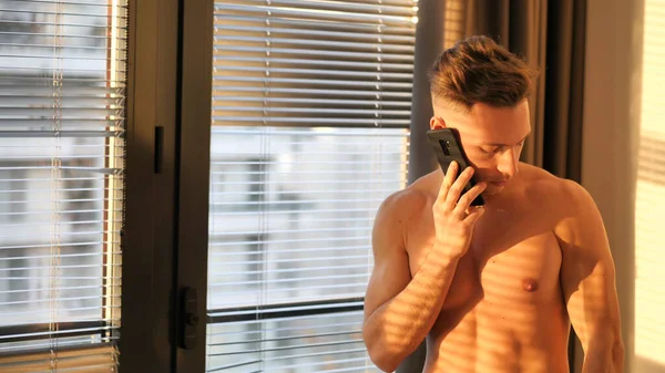 一个英俊的年轻人 光着身子 拿着一部手机 站在窗边百叶窗旁边 — 图库照片