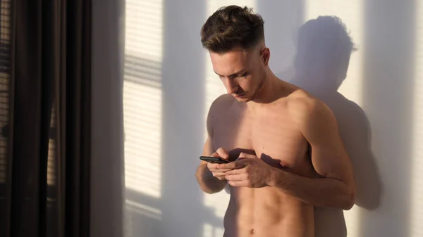 Fit handsome young man standing shirtless holding a cell phone, in his bedroom next to window blinds, typing or using it