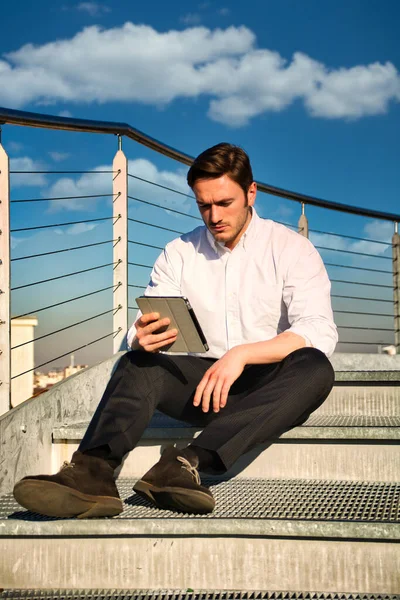 タブレットコンピュータを使って金属の階段のステップで屋外に座って白いシャツを着てハンサムな流行のビジネスマン 晴れた日に都市の設定で — ストック写真