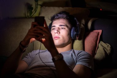 Young handsome man watching movie or listening to music on headphones, sitting on a sofa at home at night in the dark