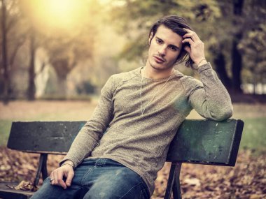 Handsome young man in city park, sitting on a bench, looking at camera, relaxing