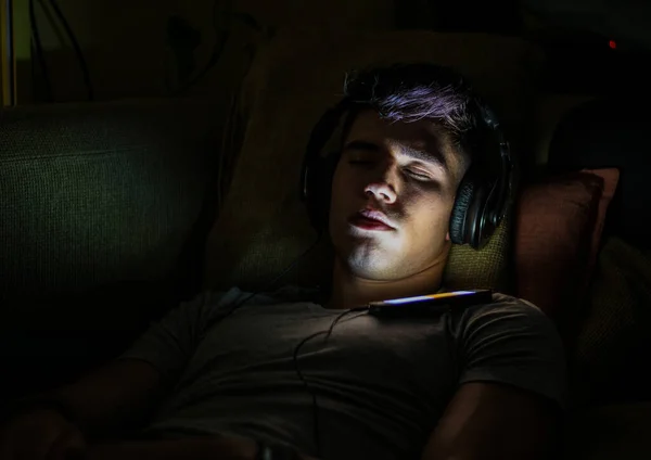 若い男性は暗い部屋でリラックスしながら携帯電話を介してプレイリストからヘッドフォンで音楽を聴く — ストック写真