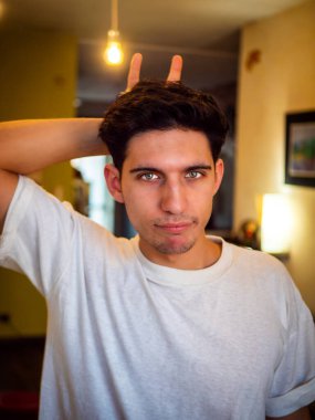 Sakin mavi gözlü genç adam boynuz hareketi yapıyor ve oturma odasında elinde ampulle kameraya bakıyor.