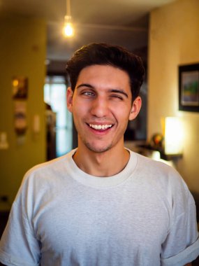 Tişörtlü, koyu renk saçlı, yanıp sönen ve evinde mutlu bir şekilde kameraya bakan adam.