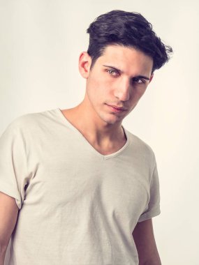 Yakışıklı ve zinde genç adam beyaz arka planda izole bir şekilde duruyor, delen yeşil gözlerle kameraya bakıyor, sade beyaz tişört giyiyor.