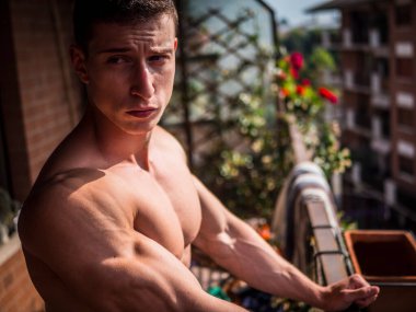 Handsome shirtless muscular young man outdoor on a balcony or terrace, wearing only jeans, looking away