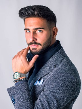 Gri arka planda, takım elbiseli ve balıkçı yaka kazaklı yakışıklı, zarif bir genç adam, kameraya bakıyor.