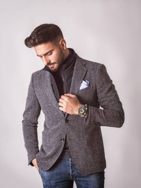 Gri arka planda, takım elbiseli, balıkçı yaka kazaklı yakışıklı, zarif bir genç adam.