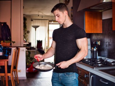 Genç erkek vücut geliştirici evde mutfakta yemek pişiriyor, nefis krepler hazırlıyor, ona bakıyor.