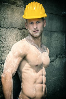Seksi, kaslı inşaat işçisi, üstsüz, açık havada çalışıyor, kot pantolon giyiyor ve sarı şapka takıyor.