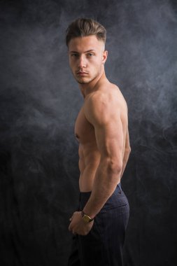 Yakışıklı, üstsüz, atletik, sadece pantolon giyen, mavi gözlü, karanlık arka planda, stüdyo çekimlerinde kameraya bakan genç bir adam.