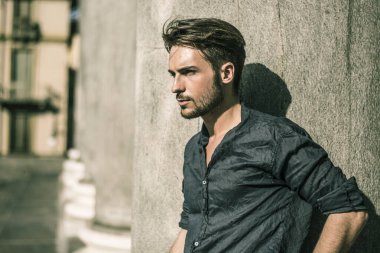 Yaz günü şehir ortamında, mavi gömlekli, taş sütun veya sütuna yaslanmış yakışıklı bir genç.