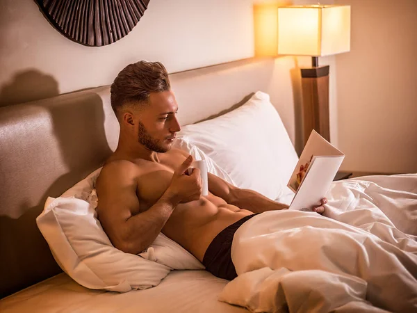 Totally naked sexy young man with muscular body on bed looking at menu or booklet or book