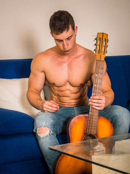 坐在沙发上弹奏古典吉他的肌肉发达的年轻人的画像 — 图库照片