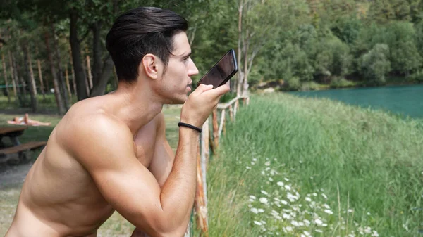 若いの上半身裸筋ハンサムな男ボイス メッセージの送信や 森の湖で スマート フォンで録音 — ストック写真