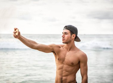 Half body shot of a handsome young man using cell phone to take photo, standing on a beach, shirtless wearing boxer shorts and baseball hat, showing muscular fit body clipart