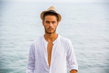 Güneşli bir yaz gününde kumsalda, şık giyinmiş, beyaz gömlek ve hasır şapka giyen yakışıklı bir genç.