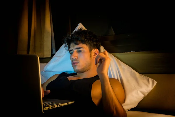 有吸引力的年轻人与严重的表情 舒适地躺在床上工作在他的创业业务的笔记本电脑 年轻的男性学院或大学生在做家庭作业 在卧室 — 图库照片