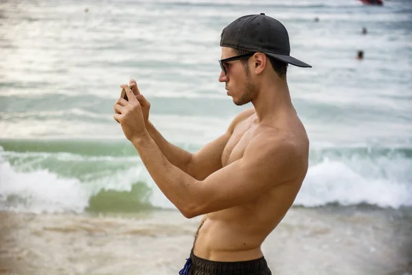 用手机拍照 站在沙滩上 赤身裸体穿着短裤 头戴棒球帽 肌肉发达的英俊年轻人的半身像 — 图库照片