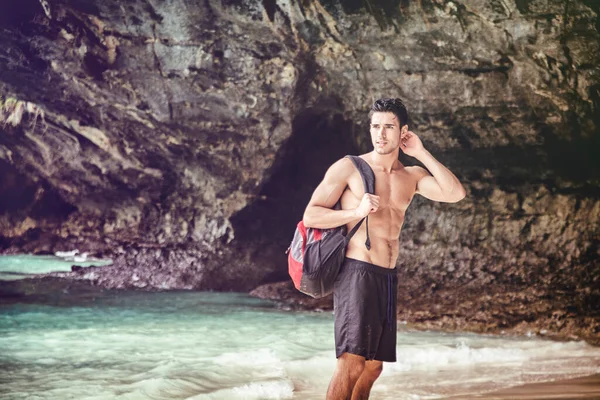 Half body shot of a handsome young man standing on a beach in Phuket Island, Thailand, shirtless wearing boxer shorts, showing muscular fit body, with backpack on one shoulder