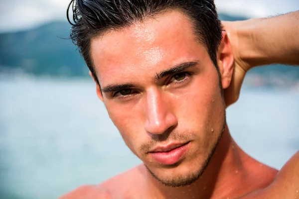 Attractive young man in the sea getting out of water with wet hair, looking at camera