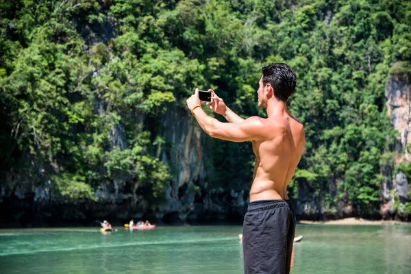 Half body shot of a handsome young man using cell phone, standing on a beach in Phuket Island, Thailand, shirtless wearing boxer shorts, showing muscular fit body