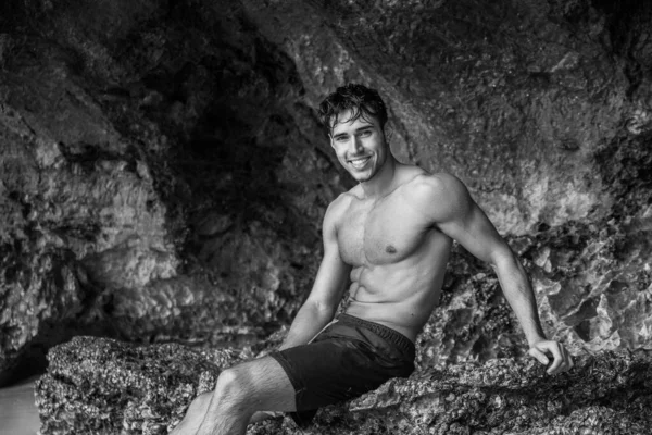 Half body shot of a handsome young man standing on a beach in Phuket Island, Thailand, shirtless wearing boxer shorts, showing muscular fit body, looking at camera in black and white shot