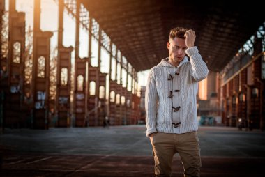 Şehir merkezinde yakışıklı bir genç, etrafında endüstriyel bir site var, kameraya bakıyor, ciddi bir ifade.