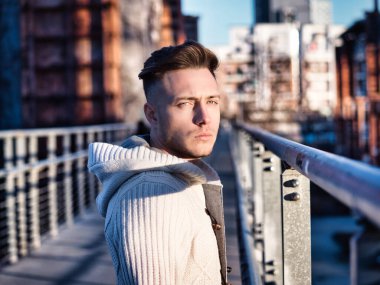 Şehir merkezinde yakışıklı bir genç, etrafında endüstriyel bir site var, kameraya bakıyor, ciddi bir ifade.