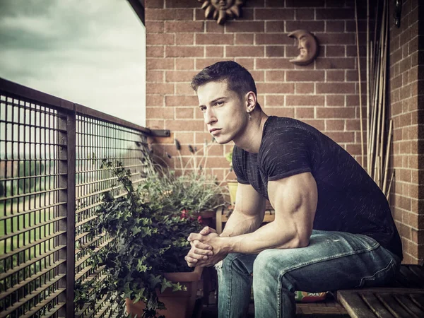 バルコニーやテラスで屋外のハンサムな筋肉質の若い男 ジーンズを着て 遠ざけ 座っている — ストック写真