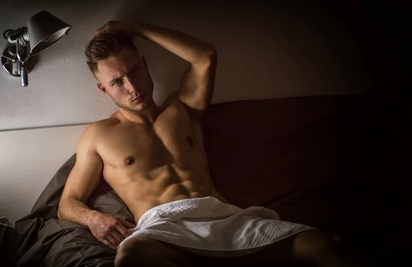 魅惑的な態度でカメラを見て 彼の寝室で彼のベッドの上だけで横になっている上半身裸のセクシーな男性モデル — ストック写真
