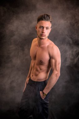 Yakışıklı, üstsüz, atletik, sadece pantolon giyen, mavi gözlü, karanlık arka planda, stüdyo çekimlerinde kameraya bakan genç bir adam.