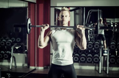 Yakışıklı genç adam, omuzlarını kaldırıyor, spor salonunda duruyor.
