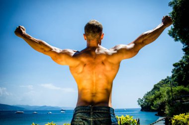 Deniz kenarında, açık havada, kaslı sırt ve omuzlarını denizin önünde gösteren genç kaslı adamın arkası.