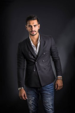 Genç işadamı kameranın önünde güvenle poz veriyor, iş elbisesini kravatsız, gömleği boynu açık, koyu arkaplanda giyiyor.