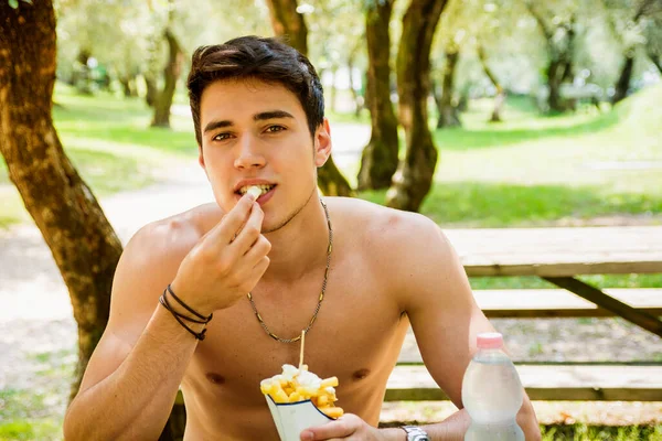 ファーストフードの昼食 ハンバーガー フライド ポテトを食べてピクニック テーブルで公園で上半身裸で座っている若い筋肉男の肖像 — ストック写真