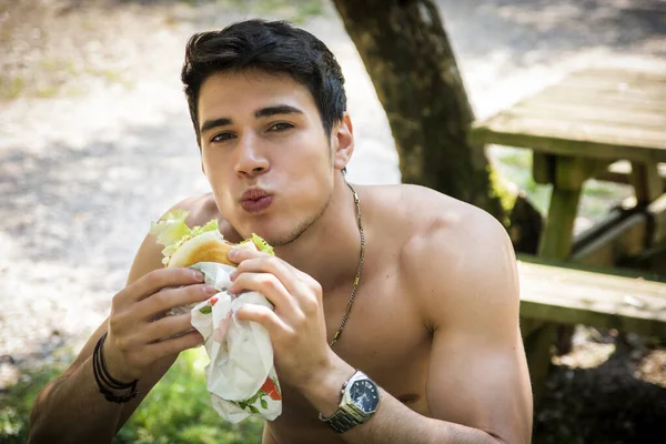 ファーストフードの昼食 ハンバーガー フライド ポテトを食べてピクニック テーブルで公園で上半身裸で座っている若い筋肉男の肖像 — ストック写真