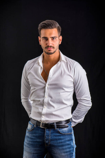 Молодой бизнесмен уверенно позирует перед камерой в белой рубашке, открытой на шее, на темном фоне