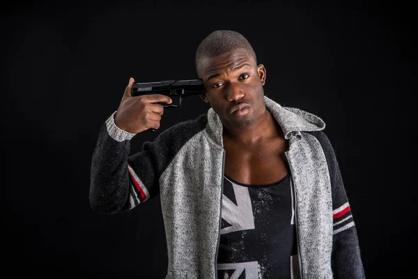 Young handsome black man holding gun at temple and looking at camera, desperate, isolated on black background