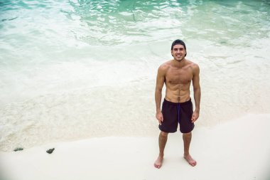 Yakışıklı bir gencin, plajda üstsüz boxer şort giyip kaslı fit bir vücut sergilediği ağır filtrelenmiş bir fotoğrafı. Tam boy çekim