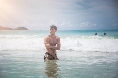 Tayland, Phuket Adası 'nda okyanusta diz çökmüş yakışıklı genç bir adam. Üstsüz, boxer şort giymiş, kaslı fit bir vücut gösteriyor.